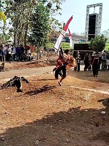 This is a short video clip of a cultural performance. In the foreground, a person in traditional clothing is dancing or performing a martial art. They are wearing a red and white patterned sarong and a striped top. They are holding a stick or a similar object. Behind them, another person is dressed in black and appears to be observing or participating. In the background, a crowd of people are watching the performance. There are trees, flags, and a stage with speakers set up. The ground is dirt and there are some hay or dried grass scattered around. The overall impression is of an outdoor community event or festival.