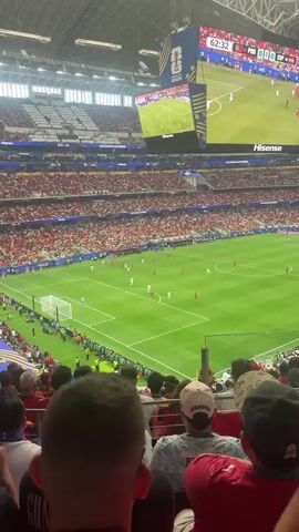The video shows a wide shot of a soccer stadium during a match. The field is green with white markings, and there are players in white and red uniforms on the field. A large screen at the top of the stadium shows the game in progress, with a score of 0-0 and the time at 62:32. The stands are filled with spectators, many of whom are wearing red. The camera is positioned in the stands, looking down at the field. The people in the foreground are wearing casual clothing, and some are wearing baseball caps. The atmosphere appears to be lively and engaged, with many spectators visible.