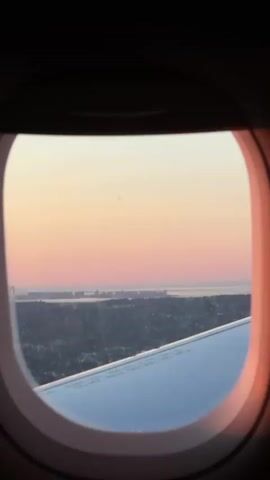 The image presents a view from an airplane window, likely captured at dusk or dawn. The dark, oval-shaped window frame encloses a landscape transitioning from land to water under a colorful sky.

In the lower right portion of the frame, a segment of the airplane's metallic wing is visible, reflecting the soft light. Some indistinct text can be faintly seen on the wing's surface. Below the wing and extending into the mid-ground, a dark landmass covered with what appears to be trees and possibly urban development stretches towards a body of water. Further in the distance, a large suspension bridge is visible spanning across the water, suggesting a significant coastal or riverine area.

The sky, which occupies the upper half of the window view, displays a gradient of soft, pastel colors. It transitions from a pale yellow or light orange at the top, blending into delicate pink and peach tones towards the horizon. The absence of clouds indicates clear weather. The overall scene suggests the aircraft is in flight, potentially ascending or descending over a populated area near Newark, United States of America, consistent with the given location context. No people are visible.