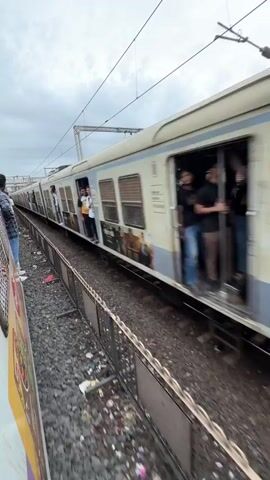 A train travels along a railway track. The train is white with blue and yellow accents and has multiple carriages. People are visible in the windows and standing in the doorways of the train. The train track is surrounded by gravel and debris. A fence runs along the side of the track. The sky is overcast.