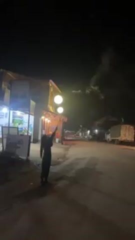 At night, an individual stands in front of a building, reaches up towards a light fixture and apparently releases a firework rocket that then rises into the night sky. A store with advertisements is seen on the left with street lights and vehicles in the background. A light fixture with two bright spherical lamps is visible above the building. A trail of light from the firework rises up into the night sky.