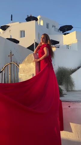 The video shows a woman wearing a long, flowing red dress with a train, posing for a photograph in an outdoor setting. She is standing near a stone pillar and a decorative metal gate, with a stark white building and a bell tower in the background. The architecture is characteristic of a Mediterranean or Greek island town, with whitewashed walls and blue-domed roofs. The sun is setting, casting a warm golden light on the scene, and the sky is a clear blue. The woman is looking towards the camera with a smile, and her dress is spread out dramatically. The camera pans slightly to capture more of the surroundings, including stairs leading up to the buildings and some dry vegetation. The overall impression is a staged photoshoot with a focus on the woman and her striking red dress against the picturesque backdrop.