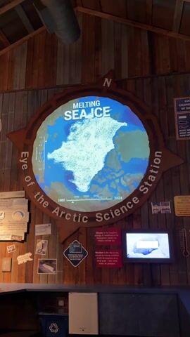 The video shows a person walking in front of a wall display with the title "Melting Sea Ice: Eye of the Arctic Science Station". The display features a digital graphic of the Arctic region, with a map showing the decrease in sea ice from 1980 to 2024. The graphic is shaped like a compass rose. There are various informational panels and photographs around the central graphic. A small video screen is also embedded in the display, showing a close-up view. The wall is made of wooden planks, and there is a recycling bin and a small refrigerator beneath the display. The overall lighting is dim, with a focus on illuminating the display.