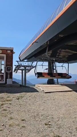 A chairlift station is shown under a bright blue sky. The station is a modern structure with a dark gray exterior and an orange trim. A single chairlift seat, with orange and black cushioning, hangs from the station, positioned over a wooden ramp. In the background, a hilly landscape stretches out under the clear sky, with some distant mountains visible. The ground in the foreground is covered with gravel and dirt. A small brick building is visible to the left of the chairlift station.
