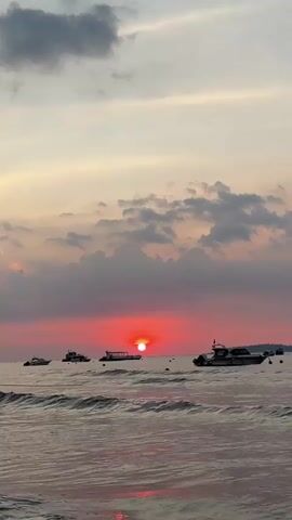 This media file captures a serene sunset over the ocean near Pemenang Timur, Indonesia. The primary subjects are several boats of various sizes floating on the water, ranging from smaller speedboats to larger vessels. The scene is set against a dramatic sky filled with a mix of light and dark clouds, which are illuminated by the vibrant, setting sun. The sun itself is a bright, fiery orb, casting a warm, reddish-pink glow across the sky and reflecting on the rippling surface of the water. The waves are gentle, creating a peaceful and tranquil atmosphere.

The time of day is clearly sunset, indicated by the low position of the sun and the intense colors in the sky. The weather appears to be partly cloudy, with the clouds adding depth and texture to the visual. Although no people are clearly visible, the presence of the boats suggests human activity related to leisure, transport, or perhaps fishing in the area. The overall mood conveyed is one of peace, beauty, and the end of a day. There is no visible text in the media file.