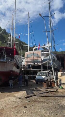 The video shows an outdoor shipyard. Several large boats are situated in a muddy area, propped up on wooden supports. One boat, named "OZEN-1," is visible with a wooden stern and a red hull. Another boat, labeled "DIVERS DELIGHT," is white with red trim and has a sign advertising scuba diving reservations. People are walking around the boats, some appearing to be working on them, and a white van is parked nearby. The sky is partly cloudy, and trees are visible in the background. Turkish flags are flown on some of the boats. A man is briefly seen pulling on a rope.