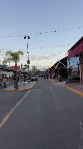 The video shows a street scene with a wide paved road in the foreground. On either side of the road are buildings with awnings, some with red fabric coverings. String lights are suspended across the street overhead. Several people are walking along the sidewalks. To the left, there is a palm tree and a street lamp. A car is parked on the left side of the street, and some benches are visible. The sky above is a pale blue with some clouds. The scene appears to be a market or a pedestrian street, possibly in the evening or during twilight hours, given the lighting.