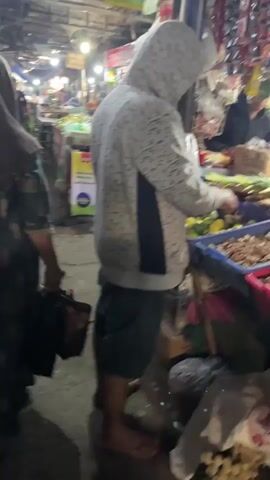 The video shows a bustling market scene. In the foreground, a person wearing a gray hooded sweatshirt with a patterned design is examining produce in a blue bin. To the right of this person, a variety of vegetables, including what appear to be limes and long, thin vegetables, are displayed. Behind this person, the market continues with stalls and more people in the background. The ground is dirty and littered with debris. Further back, another person wearing a dark patterned dress and a head covering is walking away from the camera, carrying a black bag. A motorcycle wheel is visible on the left side of the frame. The market appears to be open-air with overhead lighting and various signs and posters displayed.
