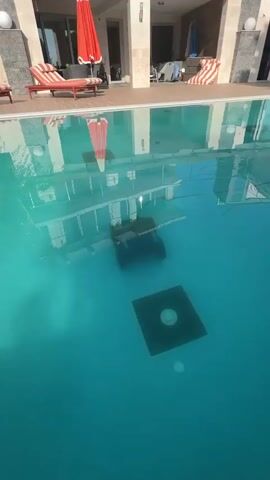 A swimming pool with clear turquoise water. At the bottom of the pool is a dark square structure with a circular fixture in the center. In the middle of the pool, a dark submerged object is visible. Above the water, reflections of a building with windows and balconies are visible. A red and white umbrella is partially submerged in the water. On the deck surrounding the pool, there are lounge chairs with red and white striped cushions.
