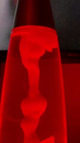 The video shows a close-up of a red lava lamp. The red wax inside is forming a bulbous shape and moving up and down within the lamp. The lamp is in front of a dark object. Audio can be heard of a man talking.