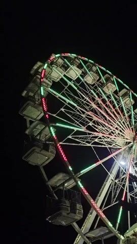The video shows a close-up shot of a Ferris wheel at night. The Ferris wheel is illuminated with red and green lights, and its cabins are visible. The background is dark, indicating that it is nighttime. The Ferris wheel appears to be in motion.