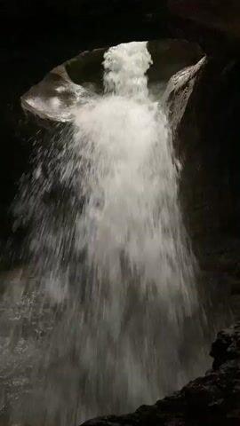 A cascading waterfall flows from an opening in a dark cave. The water appears bright white against the dark rock walls of the cave, suggesting it is well-lit by an external light source. The water plunges downwards creating a frothy spray. The rock formations around the opening of the waterfall are rough and irregular. The video is a static shot, with no movement of the camera or visible subjects other than the water.