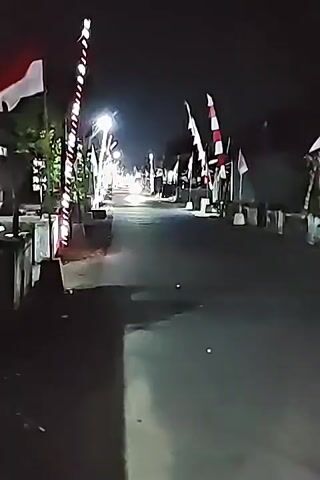 The video shows a street at night decorated with Indonesian flags. The flags are red and white, and some are attached to poles with lights. There are also decorative lights along the street. A vehicle with its headlights on is driving down the street, and it appears to be the source of the bright light in the center of the frame. The street is paved and appears wet, possibly from rain. There are buildings or structures visible on either side of the street. The scene is dimly lit overall, with the lights from the flags and the vehicle providing the primary illumination.