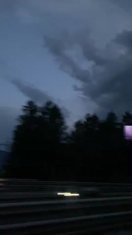 The video captures a blurred view from a moving vehicle, looking towards a dark forest line against a twilight sky with dramatic clouds. The road, marked with white lines, is visible in the foreground. A faint purple light is seen on the right side of the frame, possibly from a sign or advertisement. The overall atmosphere is dark and somber, suggesting a late evening or stormy weather.