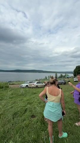 The video opens with a view of a grassy field under a cloudy sky. In the background, a body of water, likely a lake or river, stretches out, with hills or mountains on the far side. There are several cars parked near the water's edge. The foreground is dominated by tall, green grass, with a few wildflowers visible.

A woman, seen from behind, stands in the lower right corner of the frame. She has blonde hair pulled up in a bun and is wearing a light-colored tank top and a turquoise and white striped skirt or wrap. Her left hand is on her hip, and she is wearing light green Croc-style shoes. Another person, partially obscured, is standing to her left.

The camera pans slightly to the left, revealing more of the cars and the shoreline. The weather appears overcast, with dark clouds dominating the sky. The overall impression is of a casual outdoor gathering or stop by the water.