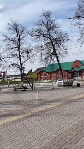 The video shows a paved public square or plaza on a sunny day. The pavement is made of grey rectangular blocks with thin yellow lines running across. There are bare trees with no leaves scattered throughout the square. A large red star monument with Cyrillic writing is visible in the middle of the square, partially obscured by metal structures that may be part of an art installation. Red flags are erected along the edges of the square. Benches and trash cans are placed along the square’s perimeter. Buildings with red and green roofs can be seen in the background across the square.  A figure on a bicycle crosses a street near the beginning of the video.