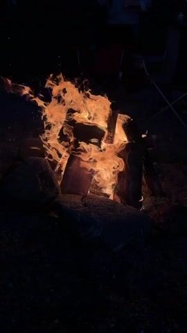 The video shows a campfire burning in the dark. The fire is contained within a ring of rocks. Flames are orange and red. Some sparks are visible. In the background, folding camp chairs and blurry figures are visible. The ground around the fire is dark and appears to be dirt or gravel.