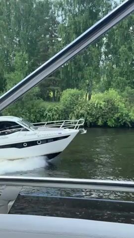 A white motor yacht speeds down a river, creating a foamy wake. The boat is a sleek, modern design with black accents along the hull and windows. The stern of the boat is visible, with an antenna on top. The yacht is surrounded by lush green trees and vegetation, indicating it is moving through a natural landscape. The water is dark and appears calm, reflecting the greenery. The camera appears to be positioned from another vessel, also likely a boat, given the close proximity and the presence of railings in the foreground. The sky is not visible, but the lighting suggests it is daytime, possibly overcast or early evening due to the soft light. The license plate on the yacht is partially visible, showing Russian characters. No people are clearly discernible on the yacht, though there may be figures inside the cabin. The overall impression is one of smooth, powerful movement through a serene natural setting.