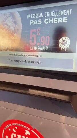 A man is shown interacting with a self-service kiosk. The screen displays "Margarita" and "Please collect your product." The man reaches into a slot below the screen and pulls out a pizza box. The box has the text "C'est qui le parrain?" and "PIZZAS GOURMETS GANG OF PIZZA" visible on it. There are what appear to be bullet holes drawn on the box. The man is looking at the screen as he retrieves the box.