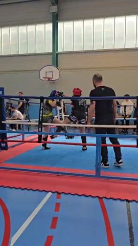 The video shows two young women engaged in a boxing match inside a ring. One woman is wearing a blue headguard and protective gear in black and yellow, while the other sports a red headguard and black and red gear. They are actively throwing punches and engaging in defensive maneuvers. A man in black clothing, possibly a coach or referee, stands near the edge of the ring. The ring is surrounded by blue and red mats on a basketball court in an indoor gymnasium. Spectators are seen sitting and watching from the sidelines. Overhead, a basketball hoop is visible, and the gym has large windows allowing natural light.