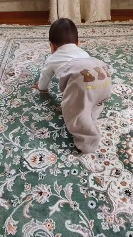 The video shows a baby crawling on a green and white patterned rug. The baby is wearing a white long-sleeved shirt and light brown pants with three teddy bear designs and the words "MOLO BALO" on the back. Another person's legs, wearing pink pants, are visible at the top of the frame, appearing to be guiding or encouraging the baby. The environment is indoors, with wooden flooring visible at the top and a curtain in the background. The camera is positioned behind and slightly above the baby, focusing on its crawling motion.