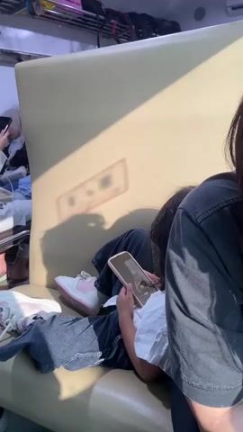 Here's a description of the video:

The video shows a young girl lying on a train seat looking at a smartphone. She is wearing jeans, a white shirt, and pink and white sneakers. An adult female, possibly her mother, sits next to her. The woman is wearing a dark gray t-shirt and glasses, and her dark hair is in a ponytail. She is also looking at a smartphone. 

Outside the train window, you can see houses and trees passing by. A bottle of water and a gray thermos are visible on a small table next to the window. The train interior appears clean and well-lit. Luggage can be seen stored on overhead racks.