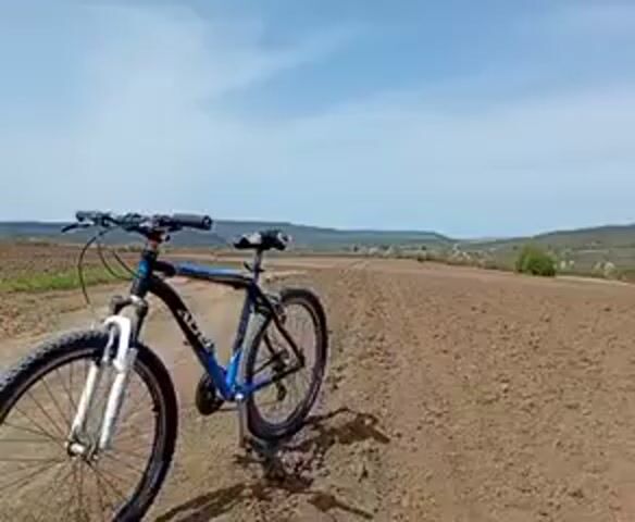 The video shows a blue and black mountain bike standing on a tilled field under a clear sky. The bicycle is positioned on the left side of the frame, with the plowed field extending towards the horizon. In the distance, rolling hills and sparse vegetation are visible. The sky is a light blue with scattered white clouds.