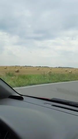 This is a first-person perspective video taken from inside a vehicle. The view is through the windshield, which shows a rural landscape. A road runs parallel to a field filled with hay bales. The sky is overcast with dramatic clouds. The interior of the car, including the dashboard, is partially visible in the foreground. The scene suggests a journey through the countryside.