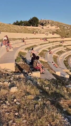 The video shows an outdoor amphitheater carved into a hillside. The seating is tiered and made of concrete, with many people seated and lounging on the steps. The sun is setting, casting a warm glow on the scene. The surrounding environment is dry and rocky, with sparse vegetation. At the center of the amphitheater, there is a circular stage with a microphone stand. The sky is clear and blue. The camera pans across the amphitheater, showing different angles of the seating and the people. Some individuals are interacting with each other, while others appear to be relaxing or enjoying the view.