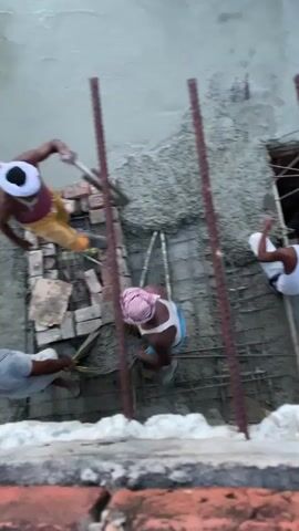 The video shows construction workers applying cement to a wall. Three men are visible, two in the foreground and one slightly to the right. The man on the left is wearing a grey shirt and is holding a trowel. The man in the center is wearing a white tank top and blue shorts and is also working with cement. The man on the right is partially visible and appears to be assisting with the construction. Rebar and bricks are present on the ground, along with some plastic piping. A yellow tarp is visible behind the workers. The scene is outdoors, and the lighting suggests it is daytime.