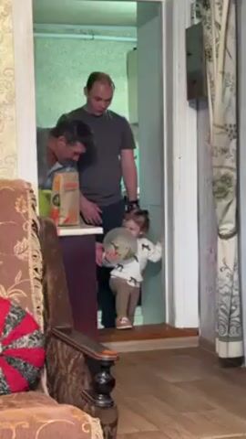 A static shot shows a toddler holding a balloon and two men standing in a doorway that leads to a kitchen. The toddler is wearing a white shirt with a horse pattern on it and brown pants and is looking down. One man is bent down and appears to be looking at something on a counter. The other man is standing up straight, also looking towards the counter. The kitchen has green tiles and a gas pipe running along the wall. The room the camera is in has a brown couch with an ornate design.