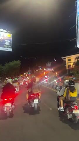 A nighttime shot from behind a motorcycle shows a busy street scene. Several motorcycles are in front, their taillights glowing red. A large billboard is visible on the left. Buildings line the right side of the street, with lights from shops and restaurants. Other vehicles, including cars and a bus, are also present. The road appears to be congested with traffic.