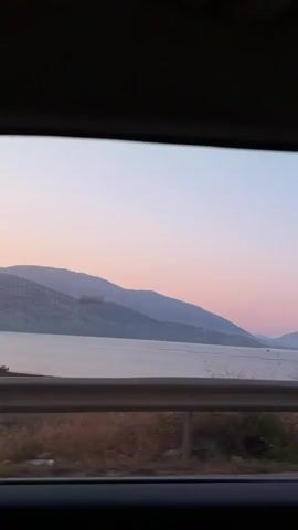 The video shows a view from inside a vehicle looking out of a window at a body of water and mountains in the distance. The sky is a soft gradient of pink and blue, suggesting either sunrise or sunset. There is a guardrail in the foreground with dry vegetation and rocks visible below it. The camera is moving, giving the impression of a scenic drive. No people or specific actions are visible, but the environment appears to be natural and tranquil.