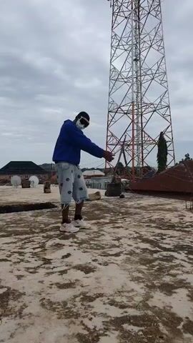 A person in a blue hoodie, denim shorts, white sneakers, and goggles is standing on a rooftop. The person is facing away from the camera and appears to be dancing or performing a ritual. A tall transmission tower is visible in the background, along with a cloudy sky. The rooftop is made of concrete and is partially covered in debris. There are also some buildings and trees in the distance.