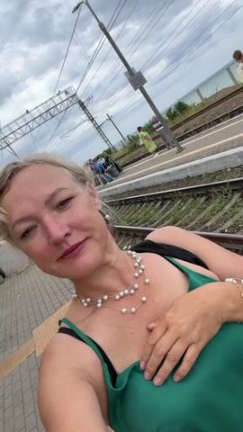 A woman is taking a selfie at a train station. She has blonde hair and is wearing a green dress, a pearl necklace, and a pearl bracelet. She is standing on a platform with train tracks behind her. There are other people and luggage visible in the background, and the sky is cloudy.
