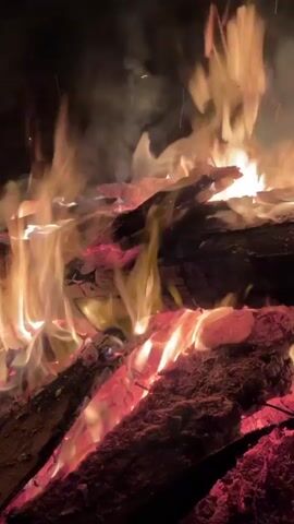 A crackling fire burns brightly in the dark, with red and orange flames enveloping large logs. Smoke rises into the black sky. The fire illuminates the wood, showcasing the char and texture. The video captures the warmth and intensity of the fire.
