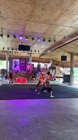 A group of men are performing what appears to be a traditional martial arts demonstration in an open-air venue. The men are dressed in what looks like historical or cultural attire, with some wearing loincloths and elaborate tops, while others are in simpler tunics and trousers. Two men are actively engaged in a mock combat, circling each other on a black mat, with one holding a sword. Another man stands nearby, also holding a sword. In the background, there are several figures, including what looks like a musician playing a stringed instrument, and individuals standing in front of draped fabric. Above the performance area, there is a thatched roof supported by wooden pillars, and a series of spotlights are directed downwards, with some casting purple and blue light onto the mat. There are also framed pictures or posters displayed on a table in the background, one of which appears to depict a person in traditional clothing. To the far left, an elephant can be seen, suggesting the cultural context of the performance. The overall environment appears to be an outdoor cultural attraction or performance space.