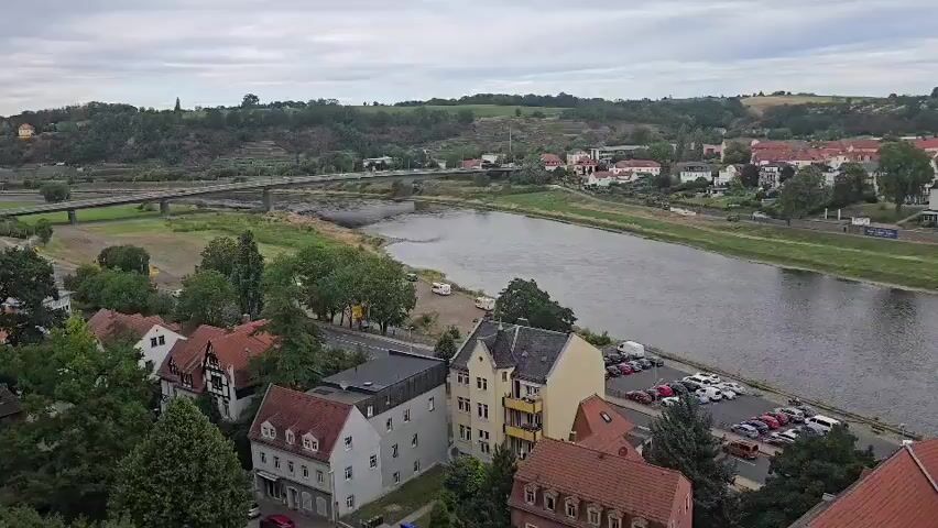 An aerial drone shot pans across a river and a town. In the foreground, there are several red-roofed buildings with detailed architecture. A church steeple is visible. Across the river, a long bridge crosses to the other side. The far bank is lined with a mixture of residential buildings and greenery. The sky is overcast.