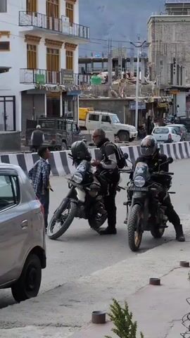 Two individuals are standing near two parked motorcycles on a street. One individual, wearing a black shirt and pants and a backpack, is looking at a mobile phone. The other individual, wearing a blue checkered shirt, is standing next to a motorcycle. A third person is seated on another motorcycle. The scene appears to be in a town with buildings and vehicles in the background.