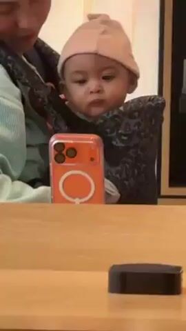 A woman is standing in what appears to be an Apple store holding a baby in a carrier. The baby is wearing a light pink beanie and looking directly at the camera with a serious expression. In front of them, on the wooden countertop, is a phone in an orange case propped up, and to the right of that, an Apple TV device. The woman is wearing a light green jacket. The lighting in the store is bright.