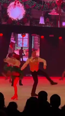 The video shows a group of ice skaters performing a Christmas-themed routine on an indoor rink. The rink is lit with red lights. A large video screen behind the skaters displays a Christmas scene of a building facade with snow falling outside the windows. The skaters are wearing Christmas-themed costumes. One female skater wears a red and green dress with a tutu, red tights, and a red hat. The male skater she is partnered with wears black pants, a white long-sleeved shirt, and a sparkly gold vest. Other skaters wear similar costumes, some also with sparkly vests. The skaters are performing a choreographed dance, skating in unison. An audience can be seen in silhouette at the bottom of the screen.