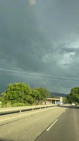 The video shows a view from a car traveling on a road. The sky is dark and cloudy, with a hint of a rainbow visible. On the right side of the road, there is a Shell gas station with a tall sign displaying fuel prices. The gas station appears to be undergoing construction or renovation, with blue barriers in front of it. Trees and greenery line the road. The camera is facing forward, capturing the road ahead.