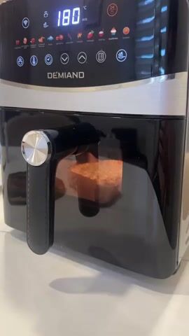The video shows a close-up of a black air fryer with a silver handle. The digital display on top shows "180" degrees Celsius. Inside the transparent drawer of the air fryer, a square-shaped food item, possibly a piece of pizza or a pastry, is visible and appears to be cooking or browning. The item has a golden-brown crust and melted cheese on top. The background is blurred but suggests a kitchen environment with white countertops.