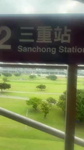 The camera looks out from behind a barrier across a wide, green field with scattered trees and walking paths. In the foreground, a sign reads "2 三重站 Sanchong Station". In the middle distance, large white letters on the grass spell out "NEW TAIPEI METROPOLITAN PARK". Several people are walking or cycling along the paths. A bridge crosses the horizon. The weather appears overcast.