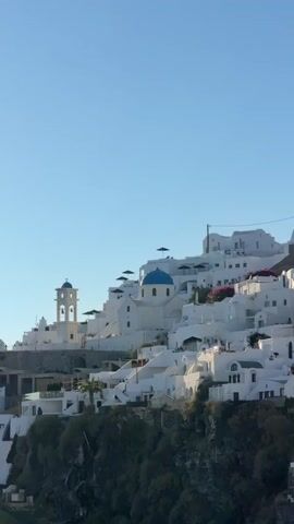 The video shows a hillside village with white buildings and blue-domed churches. The buildings are clustered together and ascend the slope, overlooking a dark green, vegetated cliff face. The sky is a clear, bright blue. The architecture is distinctly Mediterranean, characterized by whitewashed walls and a stepped layout. There are no people visible in the footage. The scene appears tranquil and bathed in natural light.
