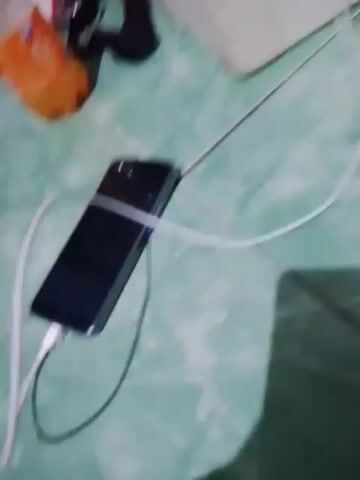 The video shows a smartphone plugged into a charger on a tiled floor. The smartphone is black and lying horizontally. A white charging cable connects the phone to a white charger plugged into an electrical outlet. There are shadows on the floor, suggesting the presence of objects and possibly a person. In the background, a bottle of orange soda and some fabric are visible.