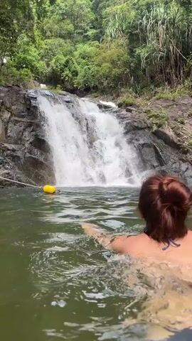 A woman is swimming in a body of water in front of a waterfall. The woman has her hair pulled back in a bun and is wearing a dark swimsuit. The water is a greenish color. There is a rope with yellow floats in the water. The waterfall is cascading down a rocky hillside covered in lush green vegetation.