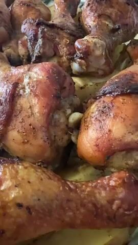 The video shows a close-up, high-angle shot of a pan filled with roasted chicken drumsticks and thighs, some of which are placed on top of sliced potatoes. The chicken pieces are golden brown and appear well-seasoned, with visible skin and some charring. The camera slowly pans across the pan, focusing on different pieces of chicken. The lighting is warm and appears to be from an overhead source, highlighting the texture and color of the food. The background is out of focus, but suggests the chicken is in a cooking pan or dish. There are no people visible in the video.