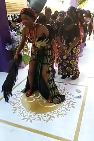 A woman dances in a wedding venue. She is wearing a strapless black dress with a high thigh slit, decorated with gold trim and red coral necklaces and headpiece. She holds a large, black feather fan in her left hand. Behind her, a line of women in matching outfits bow rhythmically. A large gold medallion design is painted on the white tile floor. Purple decorative pillars and a white tent are visible in the background.