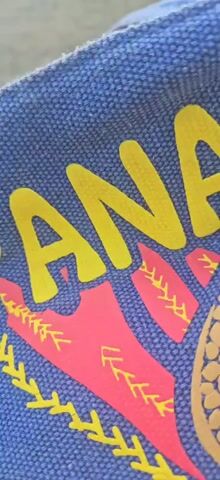 The video is a close-up shot of a blue fabric with a design on it, set against an outdoor background. The fabric appears to be a tote bag or similar item. The design features the word "PANAMA" in large yellow letters. Below the word, there is a red handprint with yellow stitching-like lines extending from it. To the right of the handprint, there is a partial image of a brown sea turtle design. The background shows a body of water, likely the ocean, and a concrete structure that could be a pier or seawall. The sky is visible with some clouds. The camera moves slightly throughout the short clip, offering different angles of the fabric and the surrounding environment. There are no people or explicit actions captured in the video.